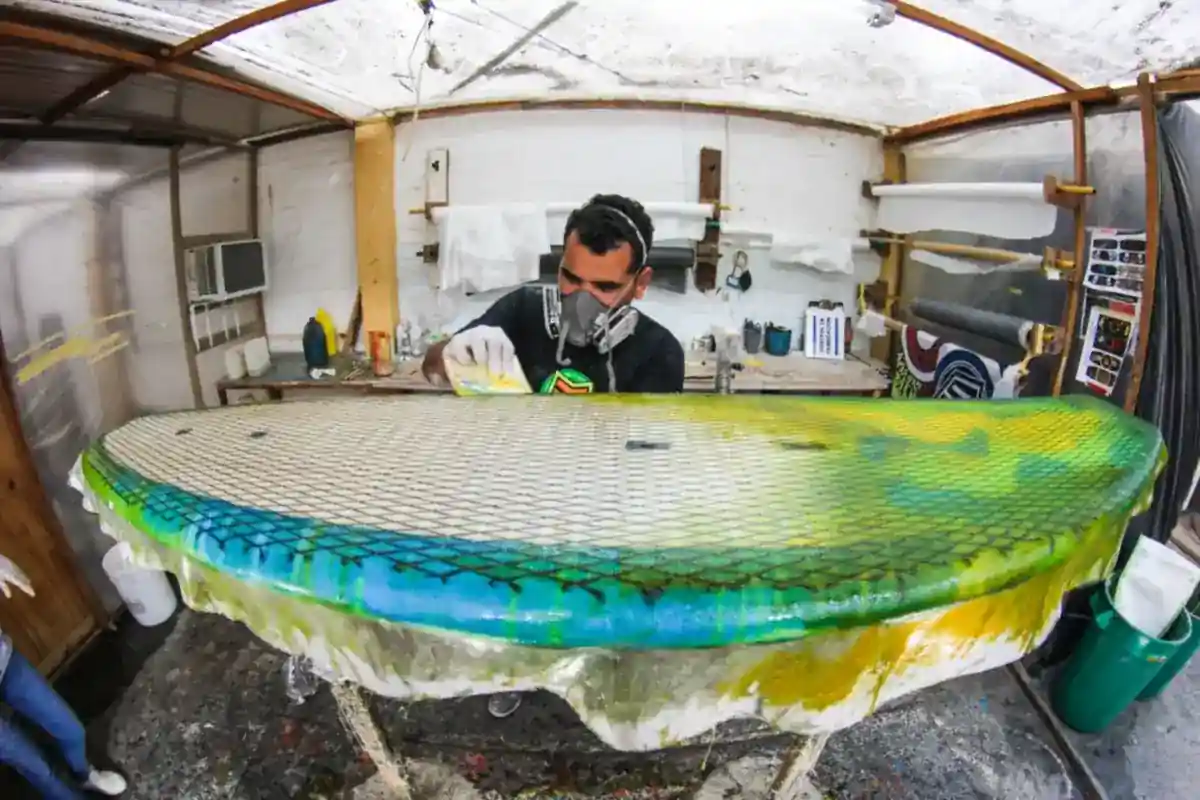 Medewerker met een ademhalingsmasker werkt aan een surfplank in een werkplaats.