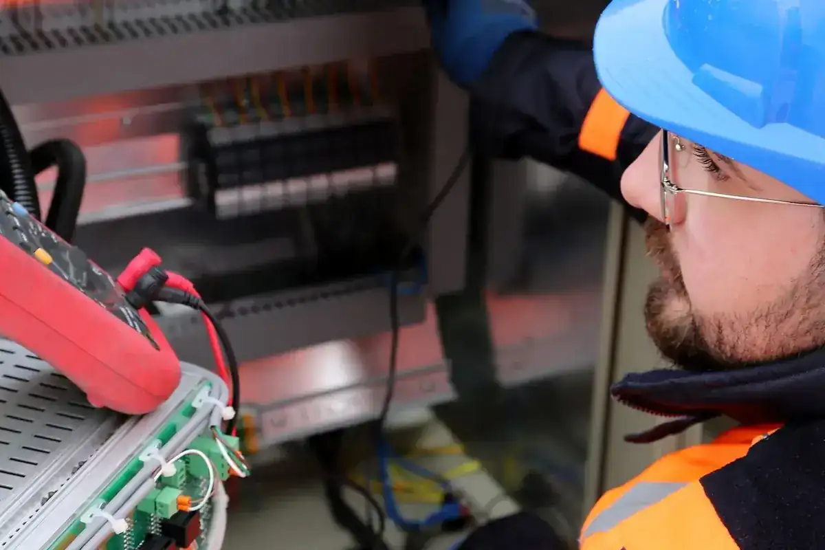 Elektricien met helm, veiligheidsbril en handschoenen aan een schakelkast.