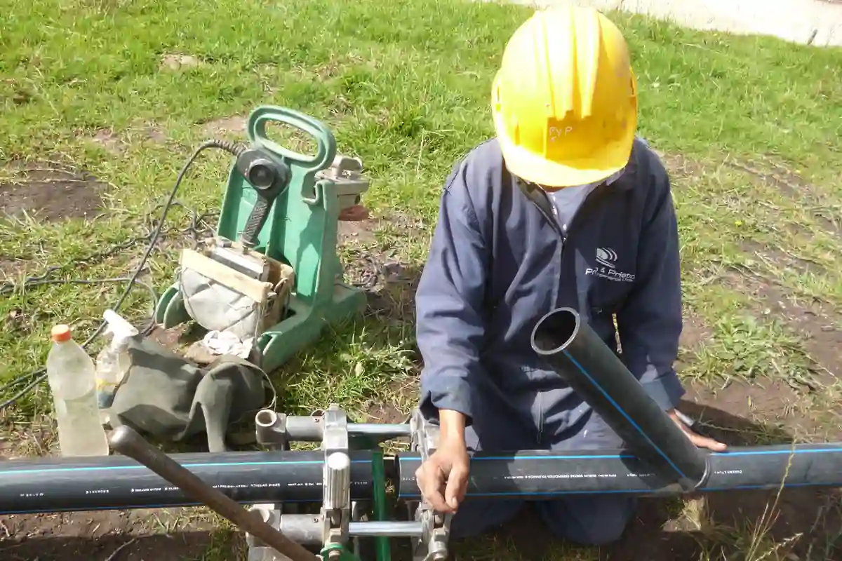 Een medewerker met helm werkt met een lasapparaat aan zwarte kunststof buizen.