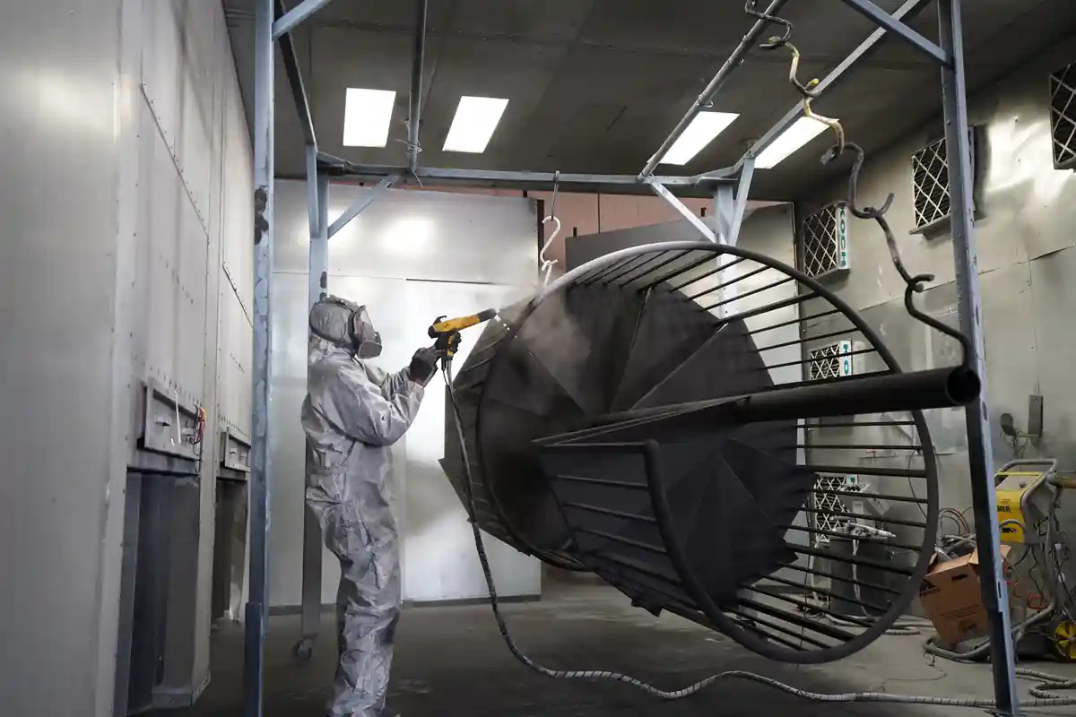 Een medewerker in beschermende kleding poedercoat een metalen constructie in een spuitcabine.