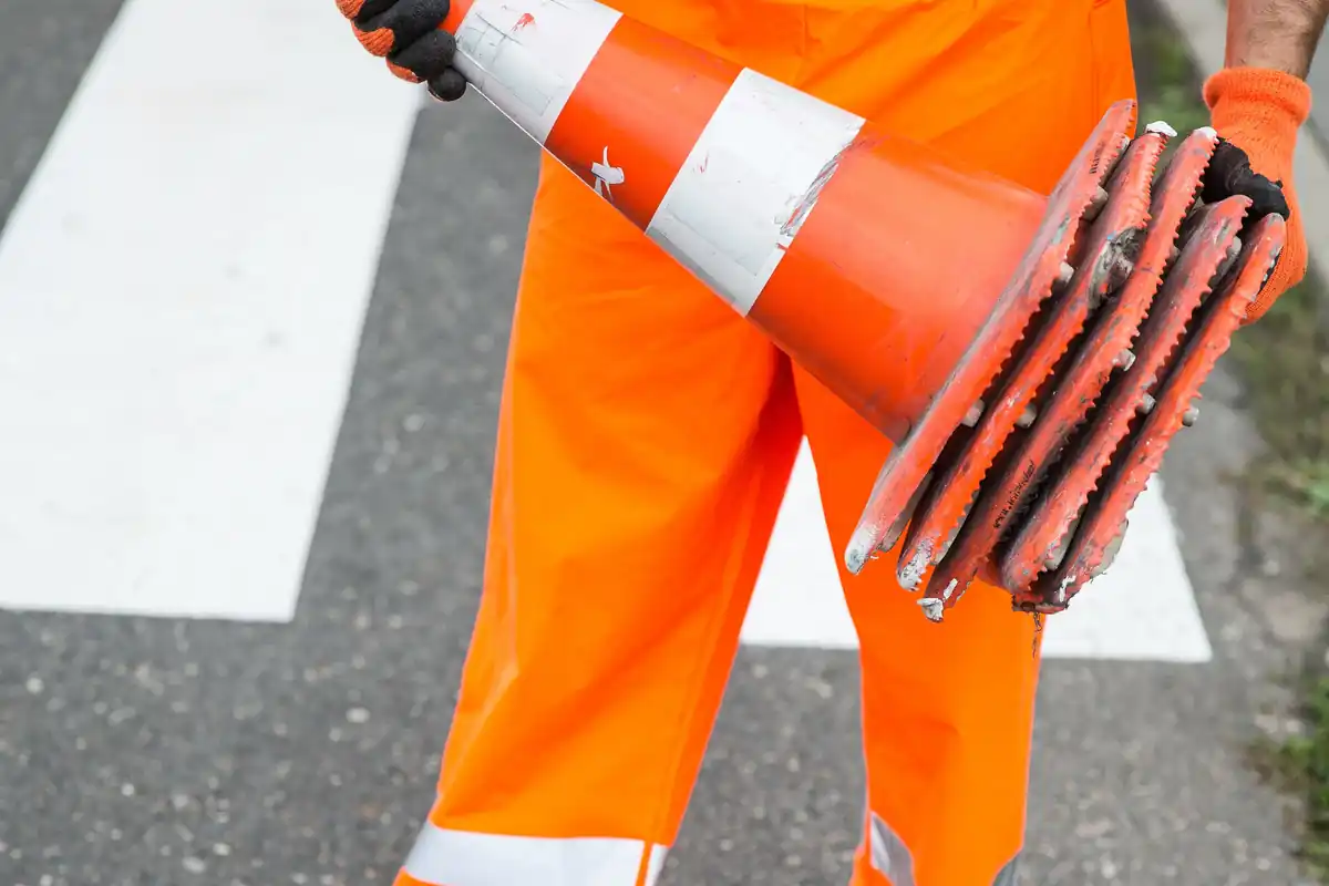 Een medewerker in reflecterende oranje werkkleding draagt een stapel verkeerskegels.